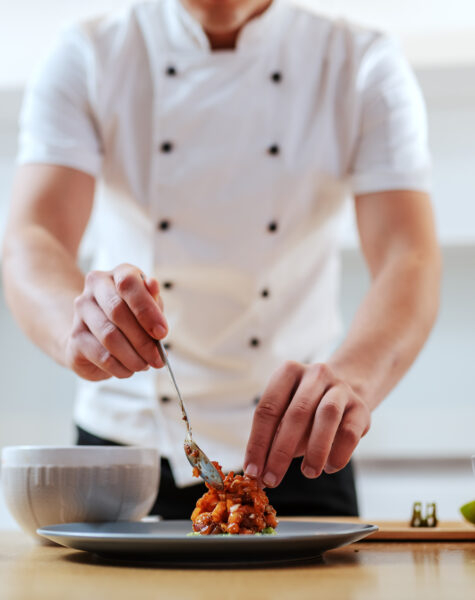 Attractive Caucasian serious chef in uniform putting prepared meal on plate. On kitchen counter are vegetables. Selective focus on hands.