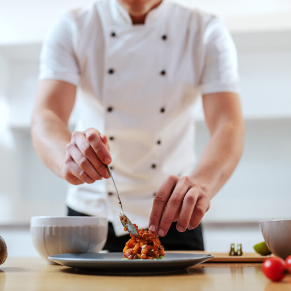 Attractive Caucasian serious chef in uniform putting prepared meal on plate. On kitchen counter are vegetables. Selective focus on hands.