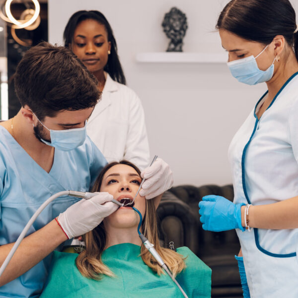 Attractive young woman in stomatology clinic being examined by a male dentist. Healthy teeth concept. Oral hygiene and dental care. Doctor fixing patient teeth with a help of a two assistant.